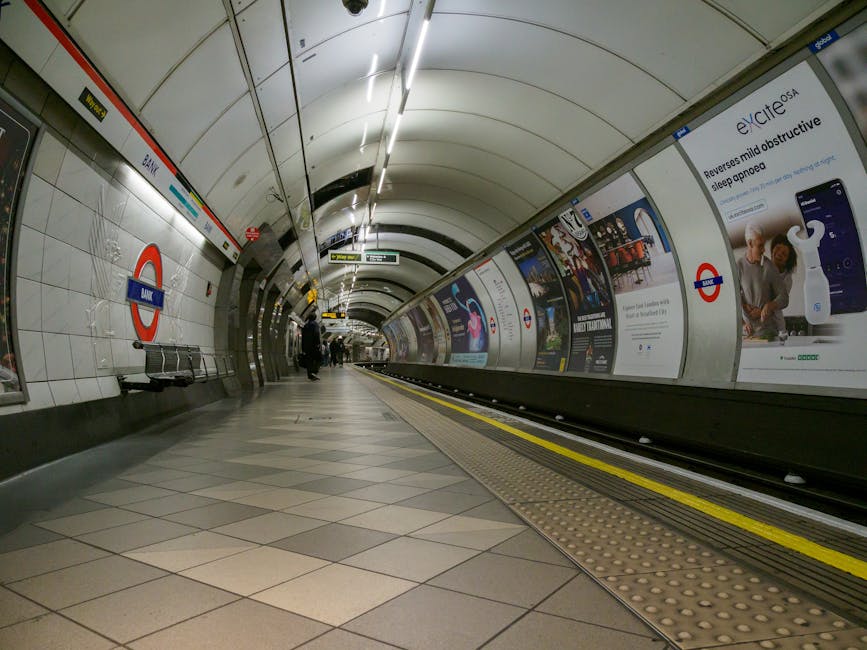 A nighttime scene at a train station platform shows a row of stationary trains on the left, with their windows illuminated, and a covered shelter running along the length of the platform on the right.