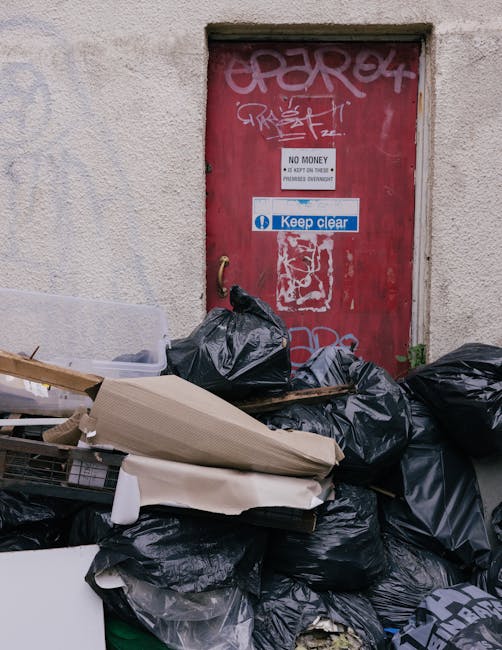 A pile of discarded household items and packaging materials situated on a dry, leafless ground surface surrounded by brown twigs and branches. The debris includes cardboard boxes, some flattened and s