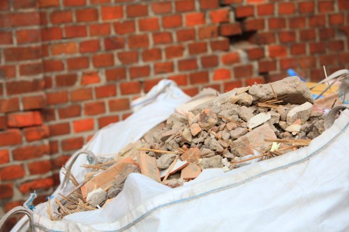 A large white construction debris bag filled with a pile of broken bricks, tiles, and concrete fragments is positioned on a paved surface next to a red brick wall. The debris includes small and medium-sized irregular pieces with rough textures and earthy tones of red, tan, and gray. Some wooden sticks and thin metal rods are partially embedded in the rubble, and a white plastic bag can be seen draped over the top of the debris in the background. The scene appears to be outdoors, possibly on a driveway or backyard area, with natural lighting highlighting the uneven surfaces and materials. This image relates to rubbish removal services offered by Garden Clearance Neasden in London, serving areas like Neasden and vicinity, with a focus on garden and construction debris clearance.