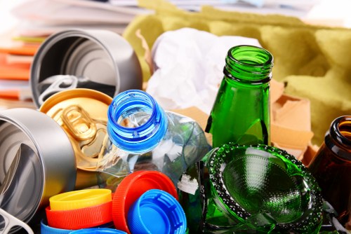 A close-up view of various recyclable waste items including clear and green glass bottles, plastic bottles with blue and yellow caps, and aluminum cans, all placed together on a surface possibly within a domestic or commercial setting. The glass bottles feature textured surfaces with some appearing smooth, reflecting light, and the plastic bottles and aluminum cans display bright colours and metallic finishes. Surrounding debris includes crumpled paper and packaging materials, with some paper visible in the background. The waste items are not contained in bins or bags but are loosely piled, suggesting a collection for rubbish removal services. The scene shows a typical assortment of recyclable rubbish that might be collected by Garden Clearance Neasden’s rubbish clearance services, situated in the Neasden area within London. Lighting in the image is natural or diffuse, illuminating the various textures and colours of the waste materials clearly. The image emphasizes the importance of waste separation and proper disposal associated with professional rubbish removal, particularly in the context of maintaining clean, clutter-free outdoor or indoor environments.